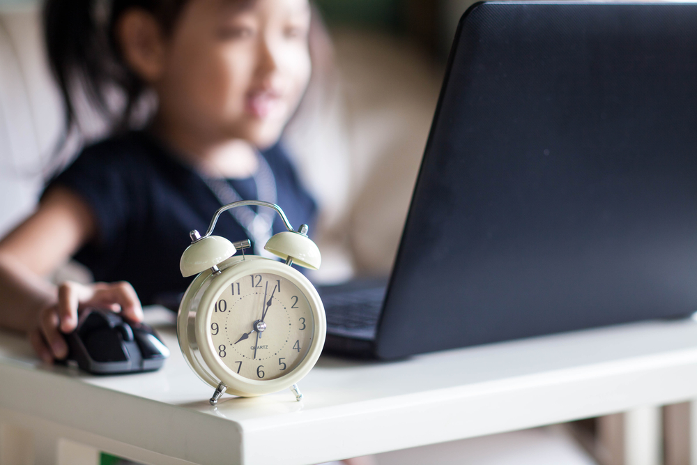 A little girl plays on the computer with an alarm clock next to it (symbolizing limited screen time)