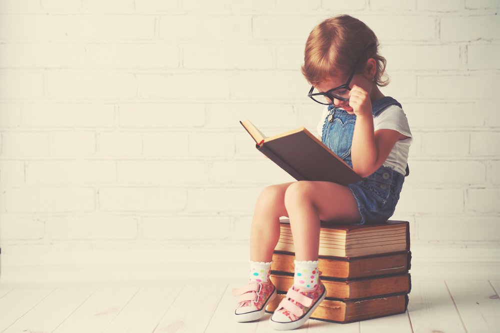 A child sits on a large stack of books reading while wearing large glasses