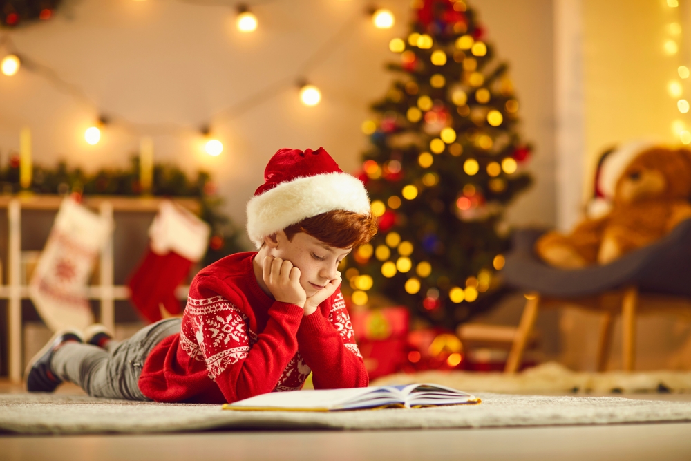Child engrossed in winter books read aloud beside a festive tree